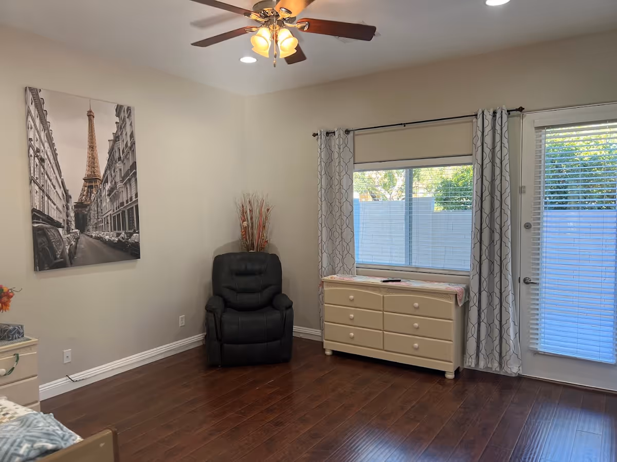 A cozy room with wooden flooring featuring a black recliner chair in the corner, a cream-colored dresser with six drawers under a window with patterned curtains, and a glass door with blinds. A ceiling fan with lights is mounted on the ceiling, and a black and white photo of the Eiffel Tower and Paris street hangs on the wall.