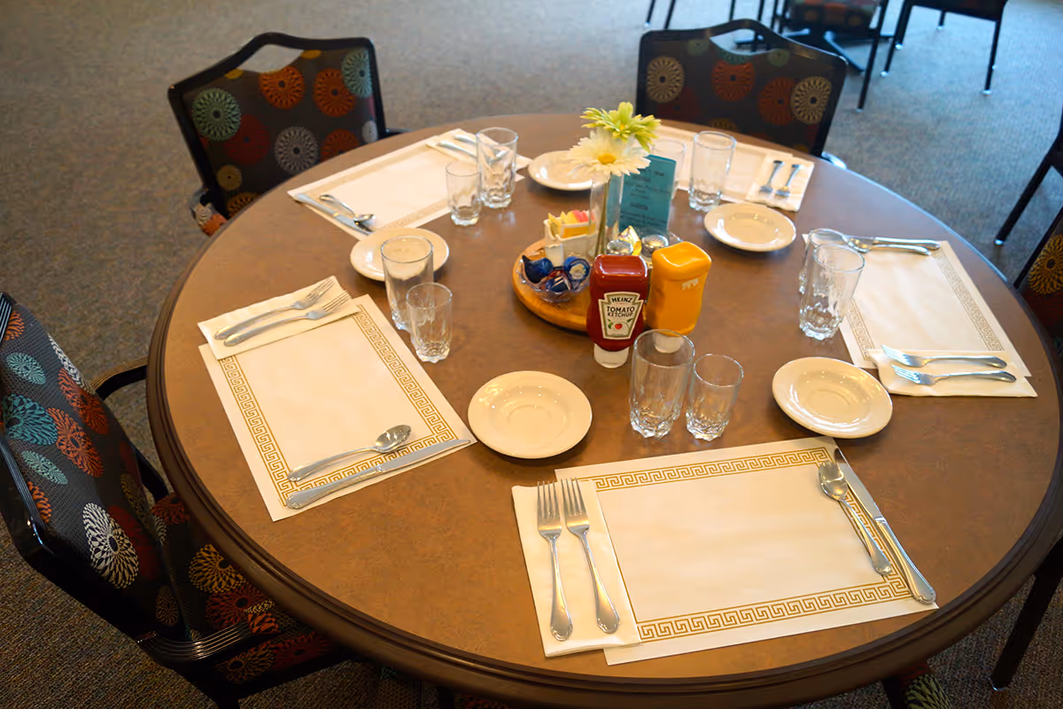 Round dining table in a communal dining room set with placemats, utensils, glasses, plates, and a condiment centerpiece.