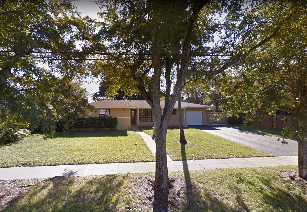 Front exterior view of a single-story residential building with a well-maintained lawn, a concrete walkway leading to the front door, a driveway, and several large trees providing shade.