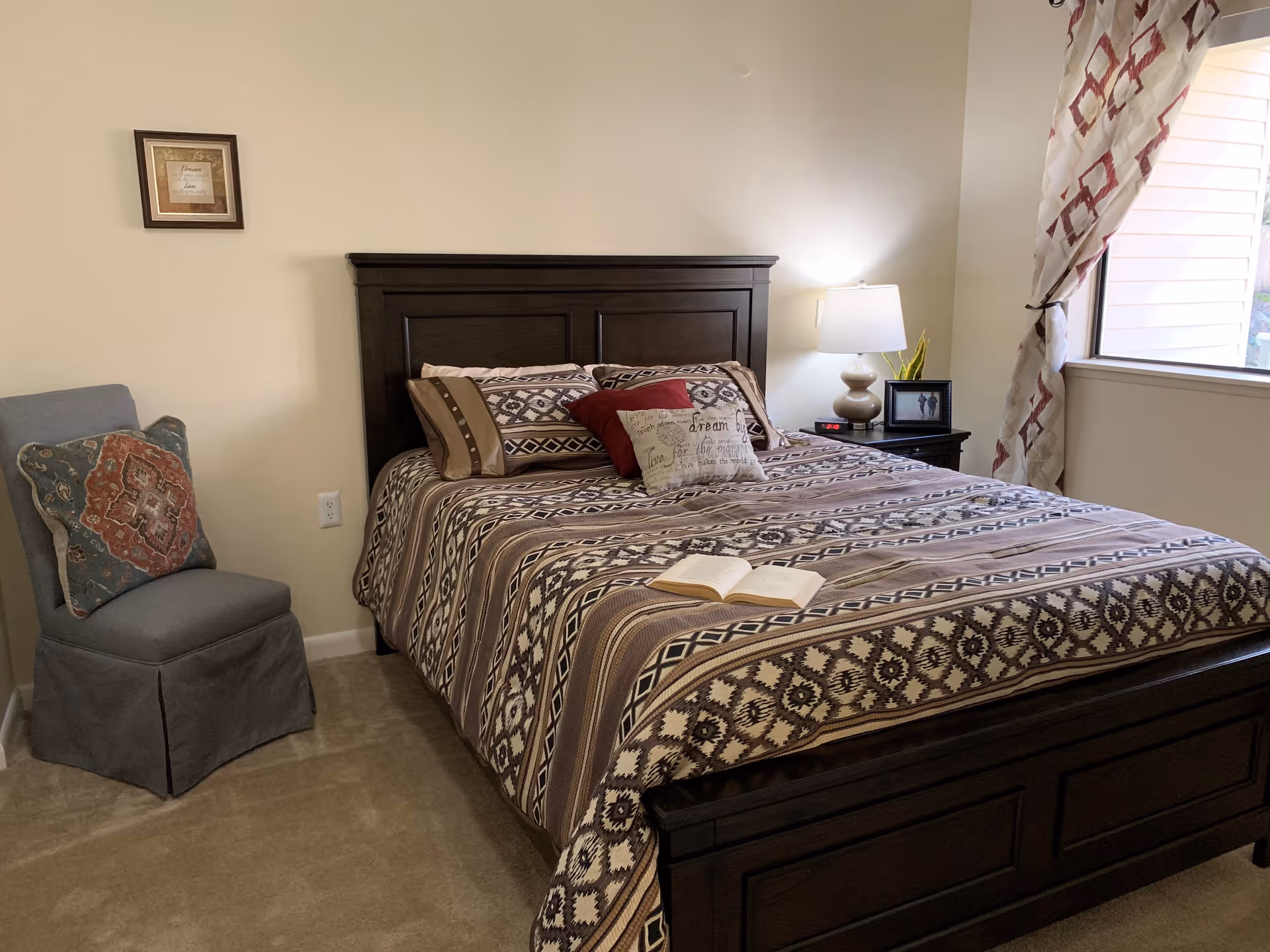 A cozy bedroom with a dark wooden bed frame and a patterned bedspread in shades of brown, beige, and cream. The bed is adorned with multiple pillows, including a red one and a decorative one with text. An open book lies on the bed. To the right of the bed is a nightstand with a lamp, a small plant, and a framed photo. A window with patterned curtains lets in natural light. On the left side of the room, there is a gray upholstered chair with a decorative pillow. A framed picture hangs on the beige wall above the chair.