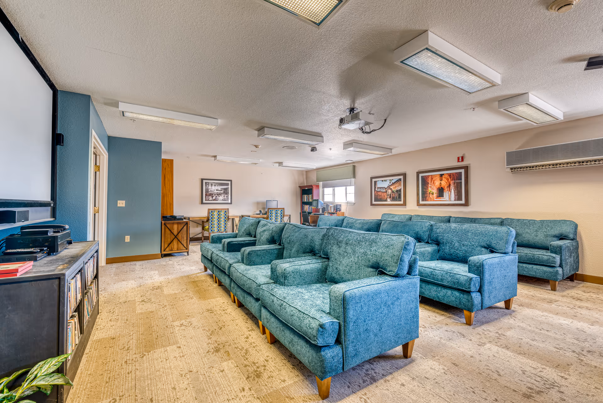A cozy common area in a senior living facility with multiple teal upholstered sofas arranged in rows facing a large screen. The room has beige walls with framed artwork, a carpeted floor, ceiling-mounted fluorescent lights, and a projector. There are also desks and chairs near the back window.