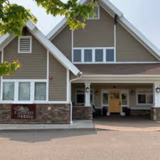 Exterior front view of a senior living facility building with a covered entrance, stone and siding facade, and a paved driveway. There are some green tree branches visible at the top left corner.