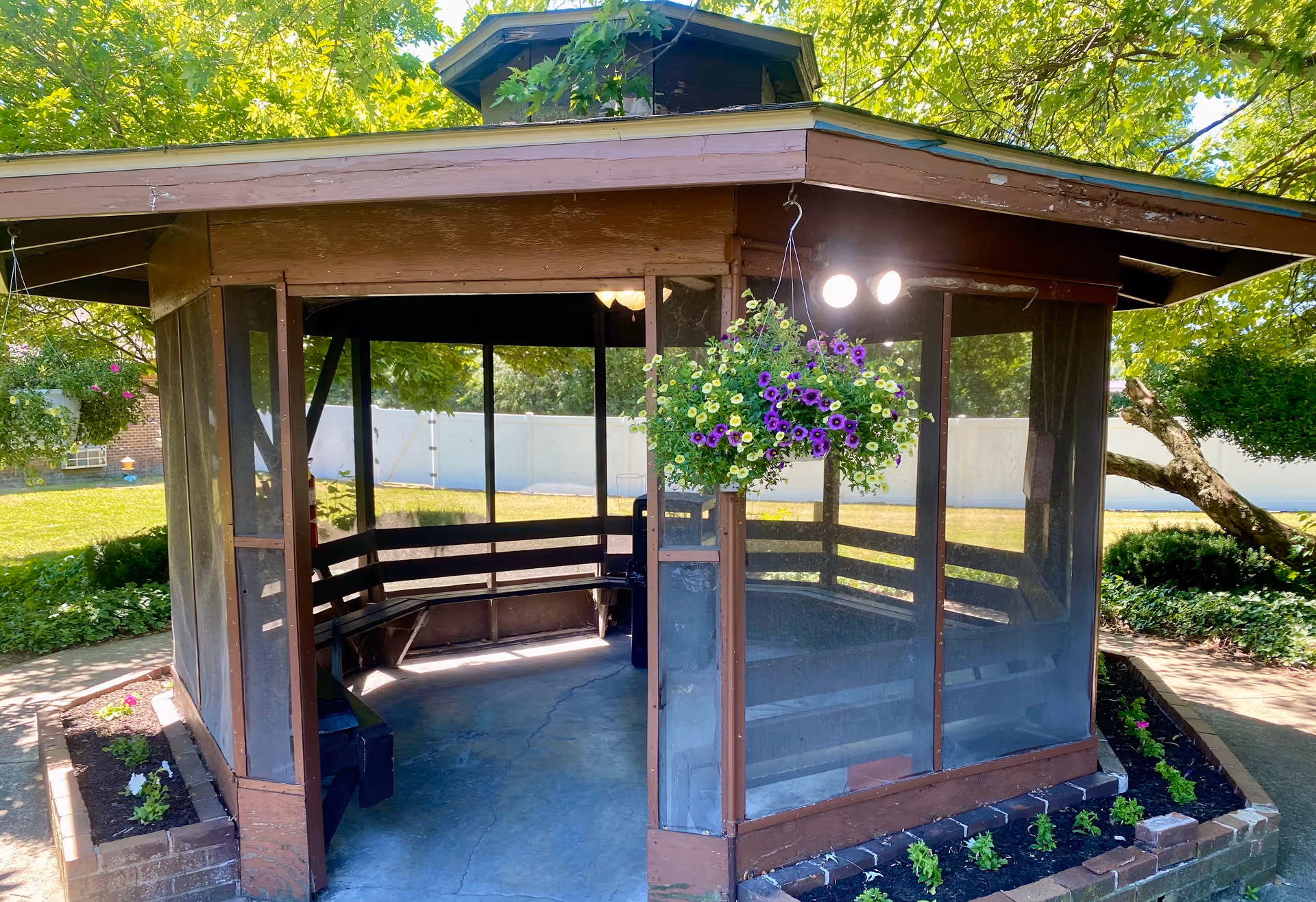 A wooden gazebo with screened walls situated in a garden area. The gazebo has built-in benches inside and hanging flower baskets with purple and yellow flowers. Surrounding the gazebo are green trees, shrubs, and a well-maintained lawn with flower beds.