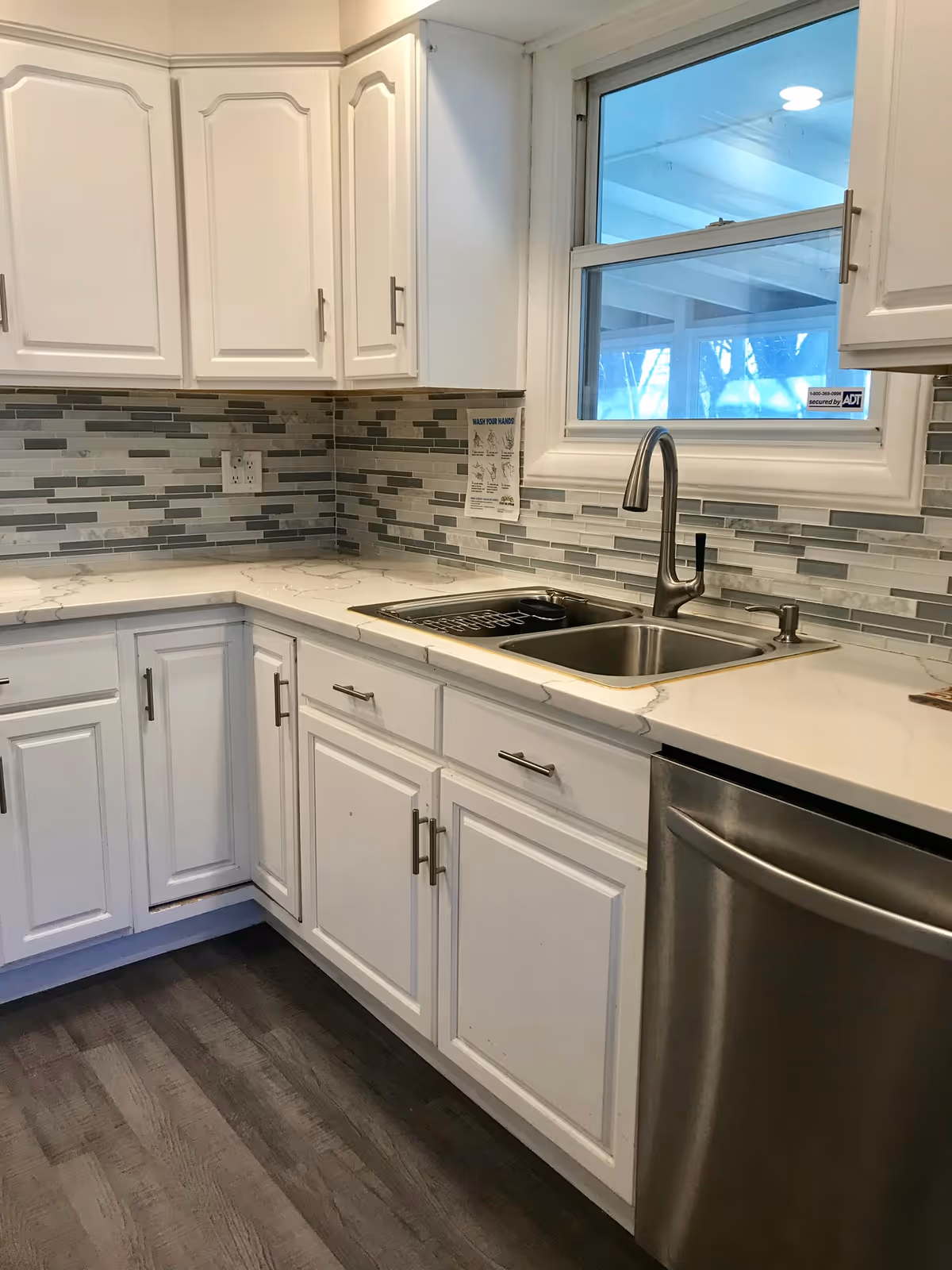 Corner of a bright kitchen with white cabinets, marble-look countertops, stainless steel sink and dishwasher, and a tiled backsplash beneath a window.