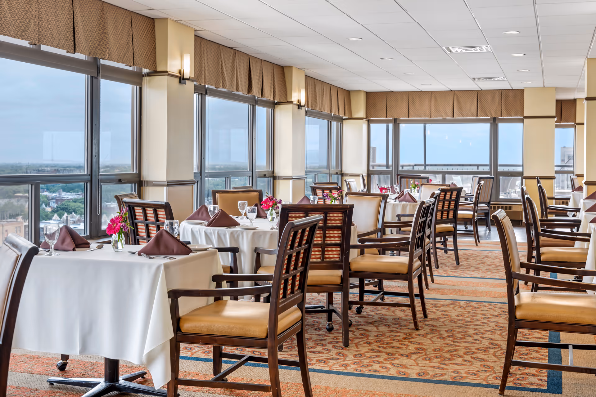 Sunlit senior living dining room with round tables, white tablecloths, and pink flowers.