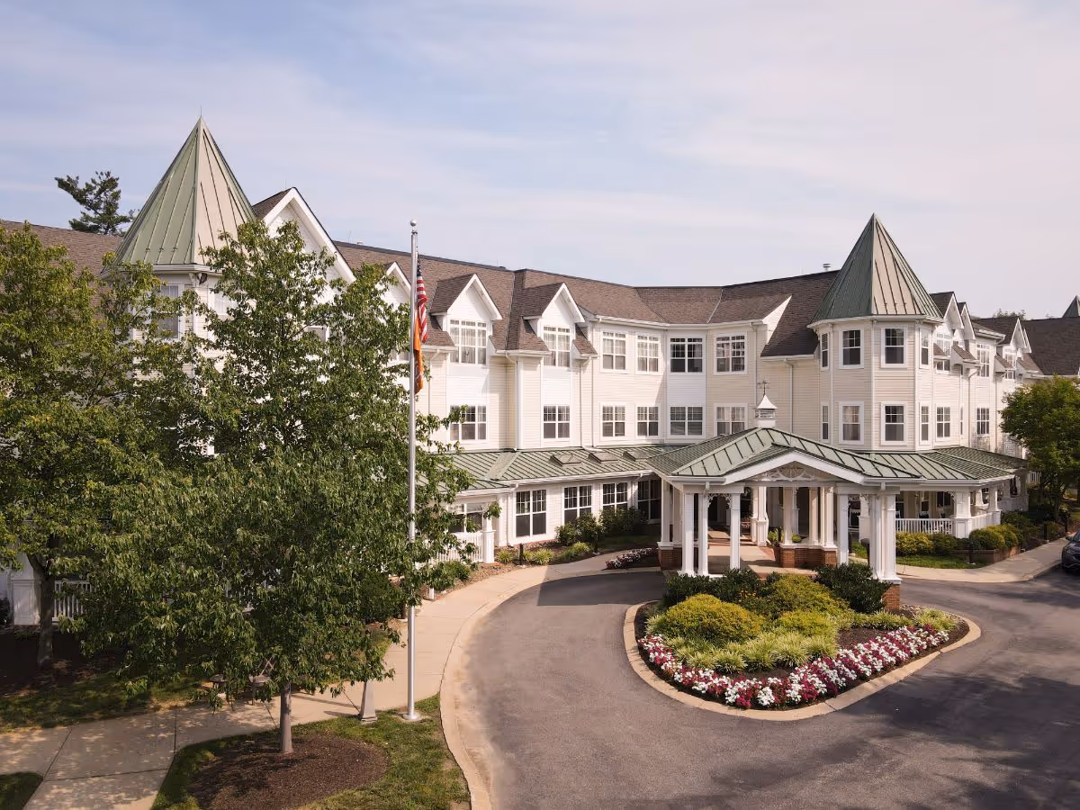 Exterior view of a large, multi-story senior living facility with white siding and green peaked roofs. The entrance features a covered driveway with white columns and a landscaped circular flower bed. Trees and a flagpole are visible near the entrance.
