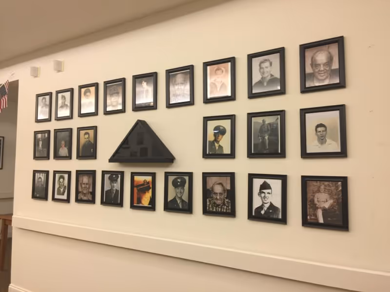 A beige wall with a collection of framed black and white and color photographs of various people, some in military uniforms, arranged in three rows. In the center of the middle row is a folded American flag in a triangular display case. An American flag is partially visible on the left side of the image.