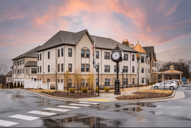 Exterior view of a three-story senior living facility building with a large decorative clock and American flag in front. The building has a combination of beige siding and stone accents, with a covered entrance and parked cars nearby. The sky is colorful with a sunset or sunrise glow.