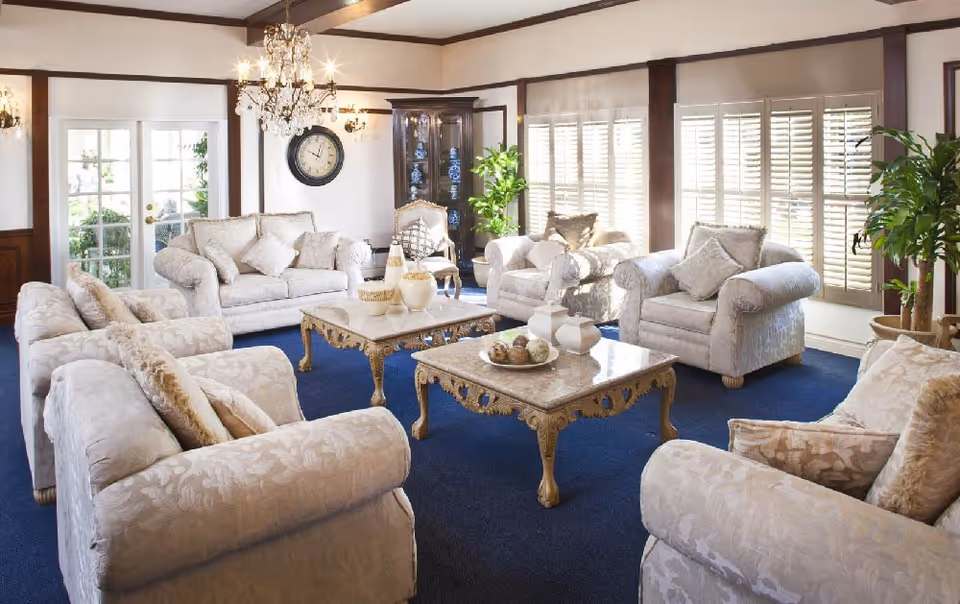 A bright and elegant living room with cream-colored upholstered sofas and armchairs arranged around two ornate coffee tables with decorative items. The room features large windows with white shutters, a blue carpet, a chandelier, wall sconces, a clock on the wall, and potted plants adding greenery.