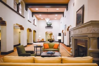Spacious and elegant living room area with high ceilings, featuring a yellow sofa in the foreground, a green armchair, and a green sofa with orange cushions around a central coffee table. The room has decorative arches along the walls, a large fireplace on the right, and warm lighting from ceiling fixtures and lamps.