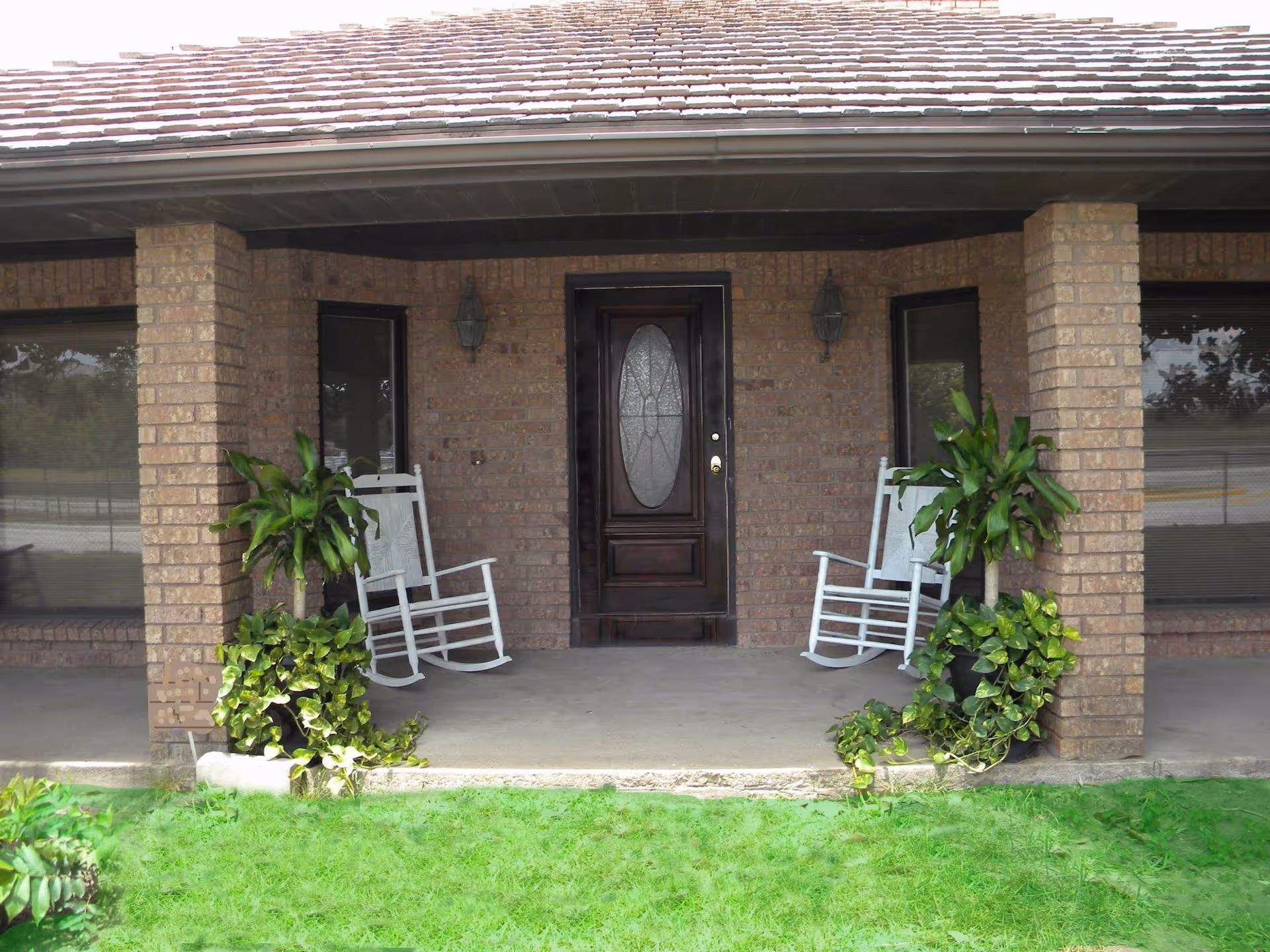 Covered brick front porch with two white rocking chairs and potted plants flanking a dark wooden front door.