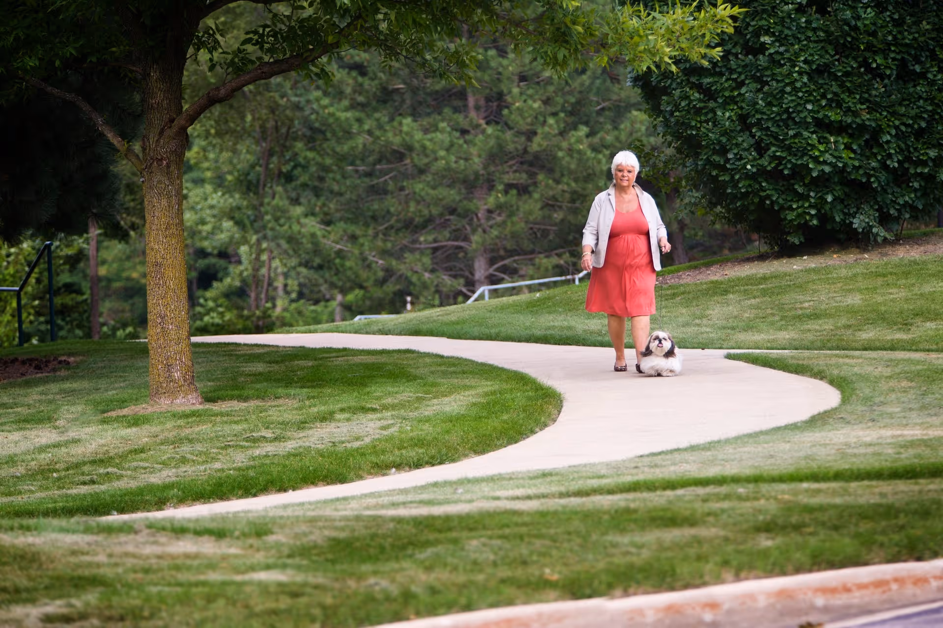 A woman in a coral dress walking a small dog on a winding paved path through a grassy, tree-lined outdoor area.