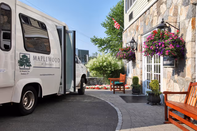 Exterior view of Maplewood at Danbury facility showing a white shuttle van parked near the entrance. The building has a stone facade with hanging flower baskets and wooden benches along the walkway. An American flag is visible above the entrance door labeled 'Main Entrance'.