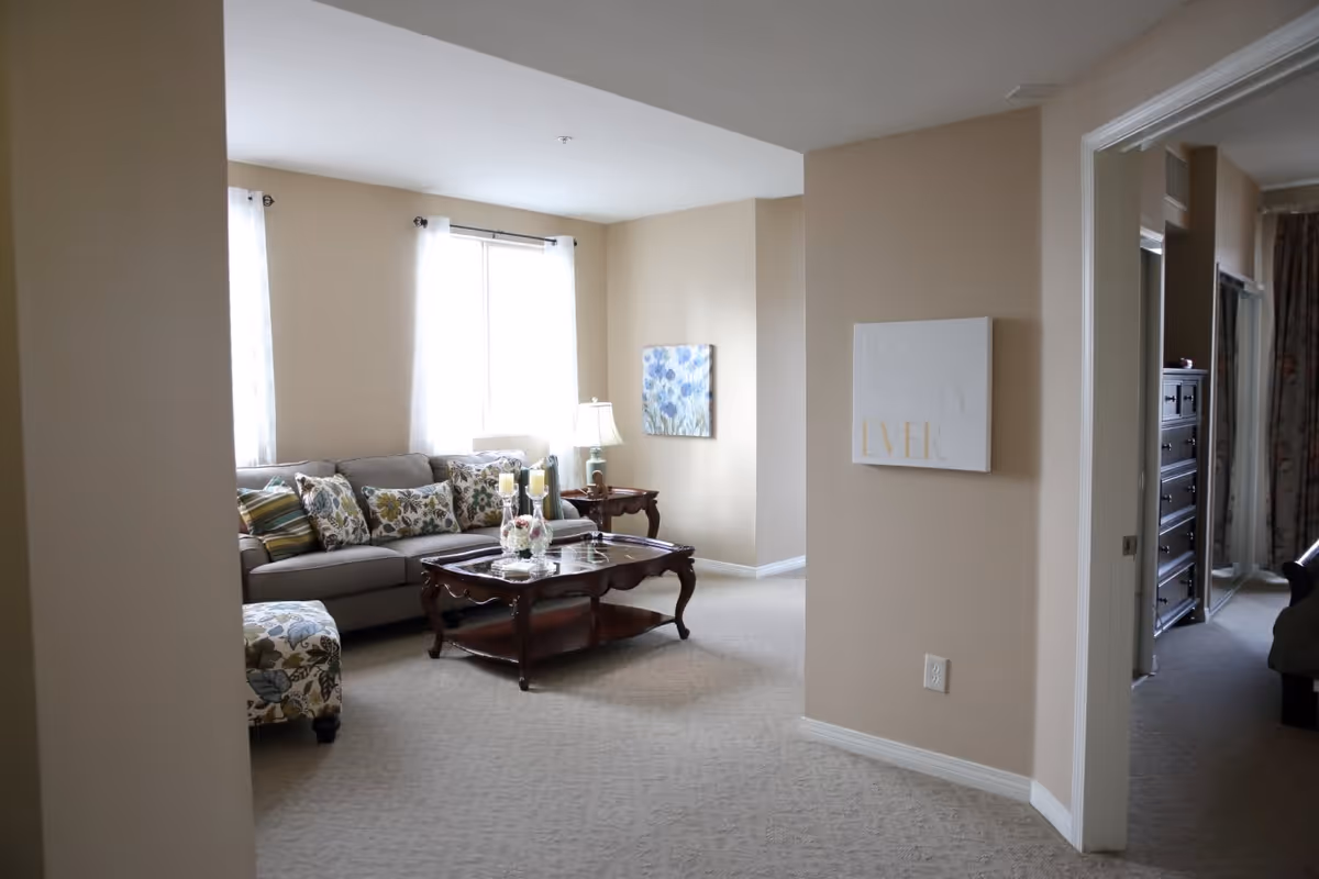 A cozy living room with beige walls and carpet, featuring a gray sofa adorned with floral and striped cushions, a wooden coffee table with decorative items, a matching floral ottoman, and a side table with a lamp. Two windows with sheer white curtains allow natural light to fill the room. A hallway leads to another room with a dresser and curtains visible.
