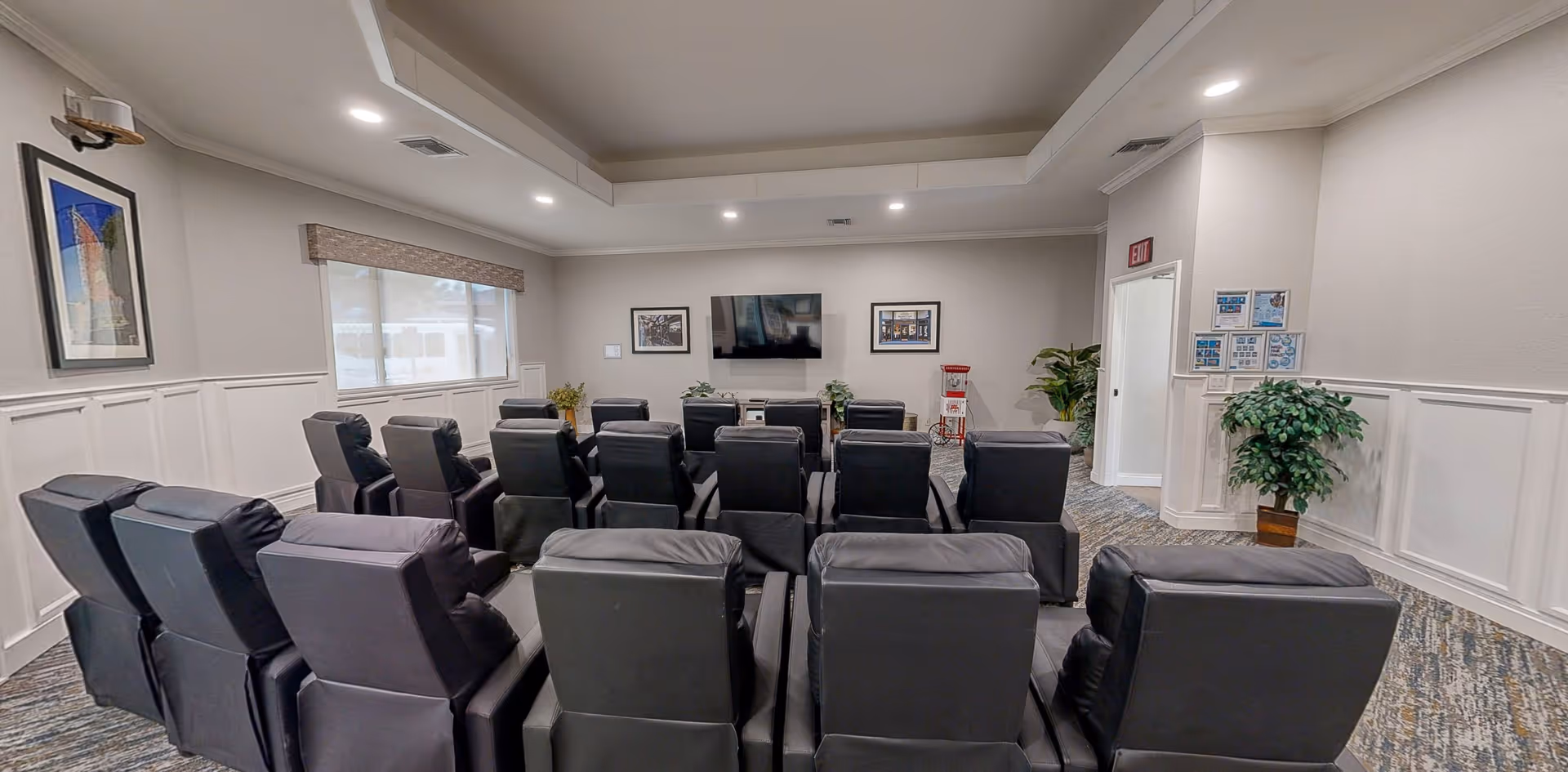 A small theater-style room with three rows of black leather recliner chairs facing a wall-mounted flat screen TV. The room has light-colored walls with white wainscoting, a window with a blind, framed pictures on the walls, and two potted plants. There is an exit door on the right side and a popcorn machine near the TV.