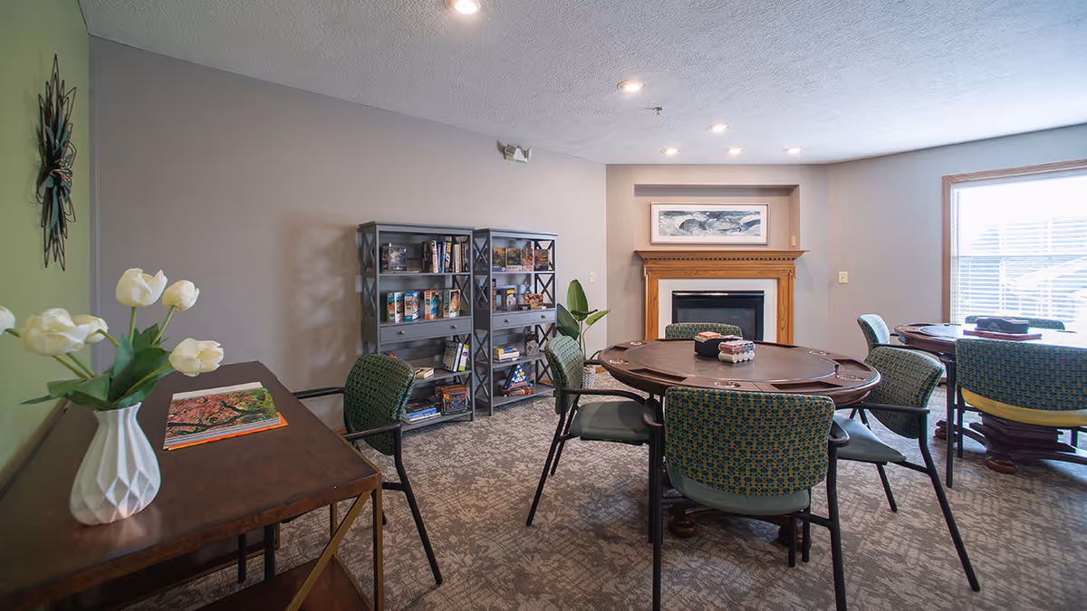 A cozy common area with round tables surrounded by chairs, a fireplace with a framed picture above it, a bookshelf filled with books and games, and a table with a vase of white flowers and a book on it. The room has carpeted flooring and large windows letting in natural light.