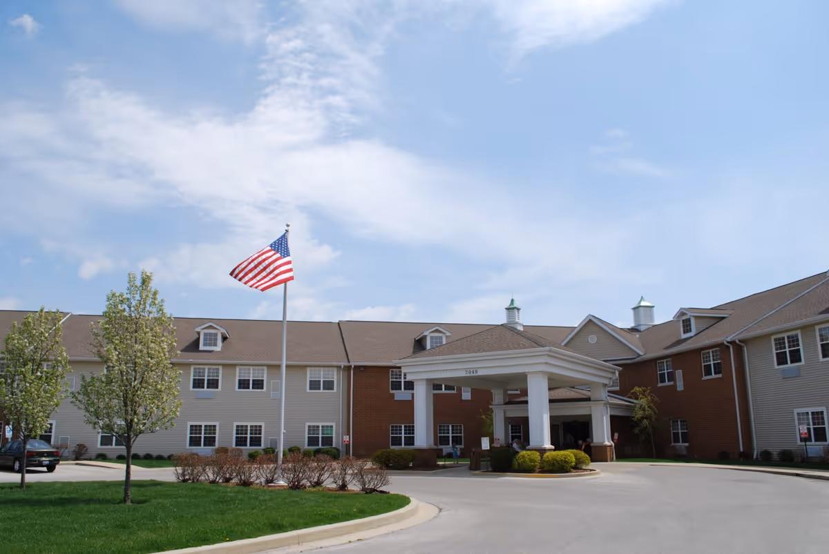 Exterior view of Heritage Woods Of Centralia senior living facility showing a large two-story building with beige and brick siding, a covered entrance, an American flag on a flagpole, and a driveway surrounded by green grass and trees.