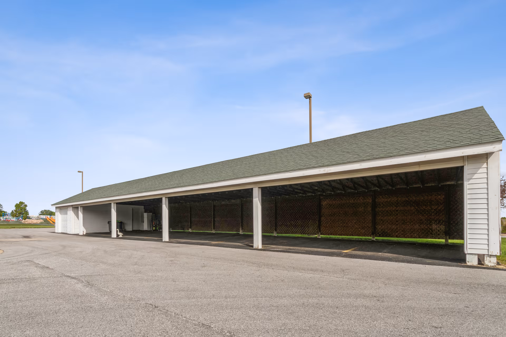 Long open-sided covered parking/carport with a pitched roof in an empty paved lot under a clear blue sky.