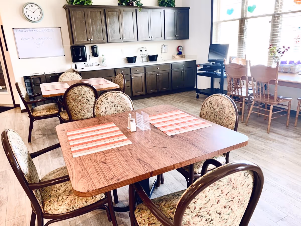 A bright dining area in an assisted living facility with wooden tables and floral upholstered chairs. The tables have red and white striped placemats, salt and pepper shakers, and a clear plastic holder. In the background, there are dark wooden cabinets with a countertop, a wall clock, a whiteboard with a message, and a window with wooden chairs and plants on the windowsill.