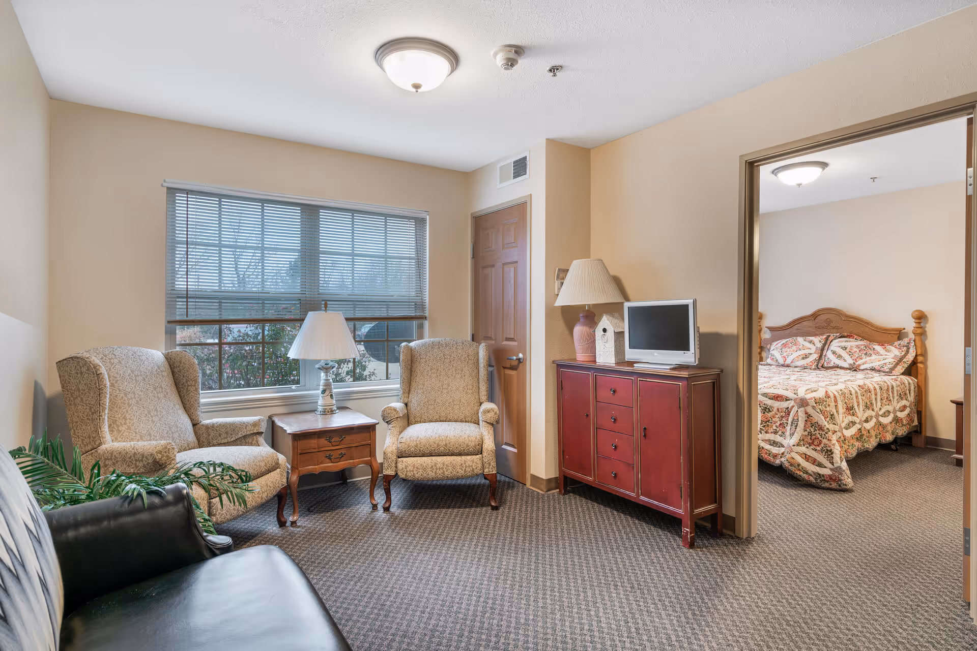 Cozy sitting area with two upholstered armchairs, a side table and lamp, a red cabinet with TV, and a view into an adjacent bedroom.