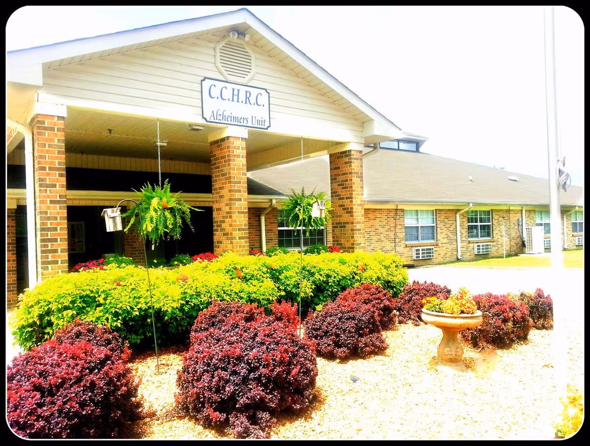 Exterior view of Cherokee County Health and Rehabilitation Center's Alzheimer's Unit entrance with brick columns, a covered porch, and well-maintained landscaping including green and red bushes and hanging plants.