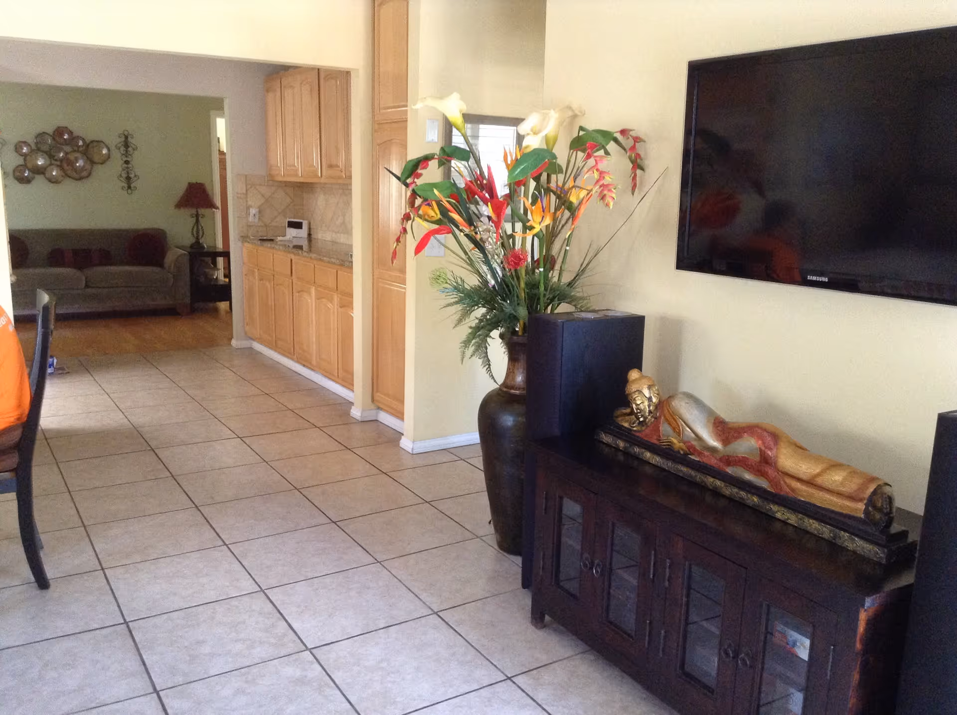 Interior view of a living space with tiled floor, a large vase with colorful flowers, a wooden cabinet with a reclining Buddha statue on top, a mounted flat-screen TV, and a glimpse into a living room area with a sofa and wall decorations.