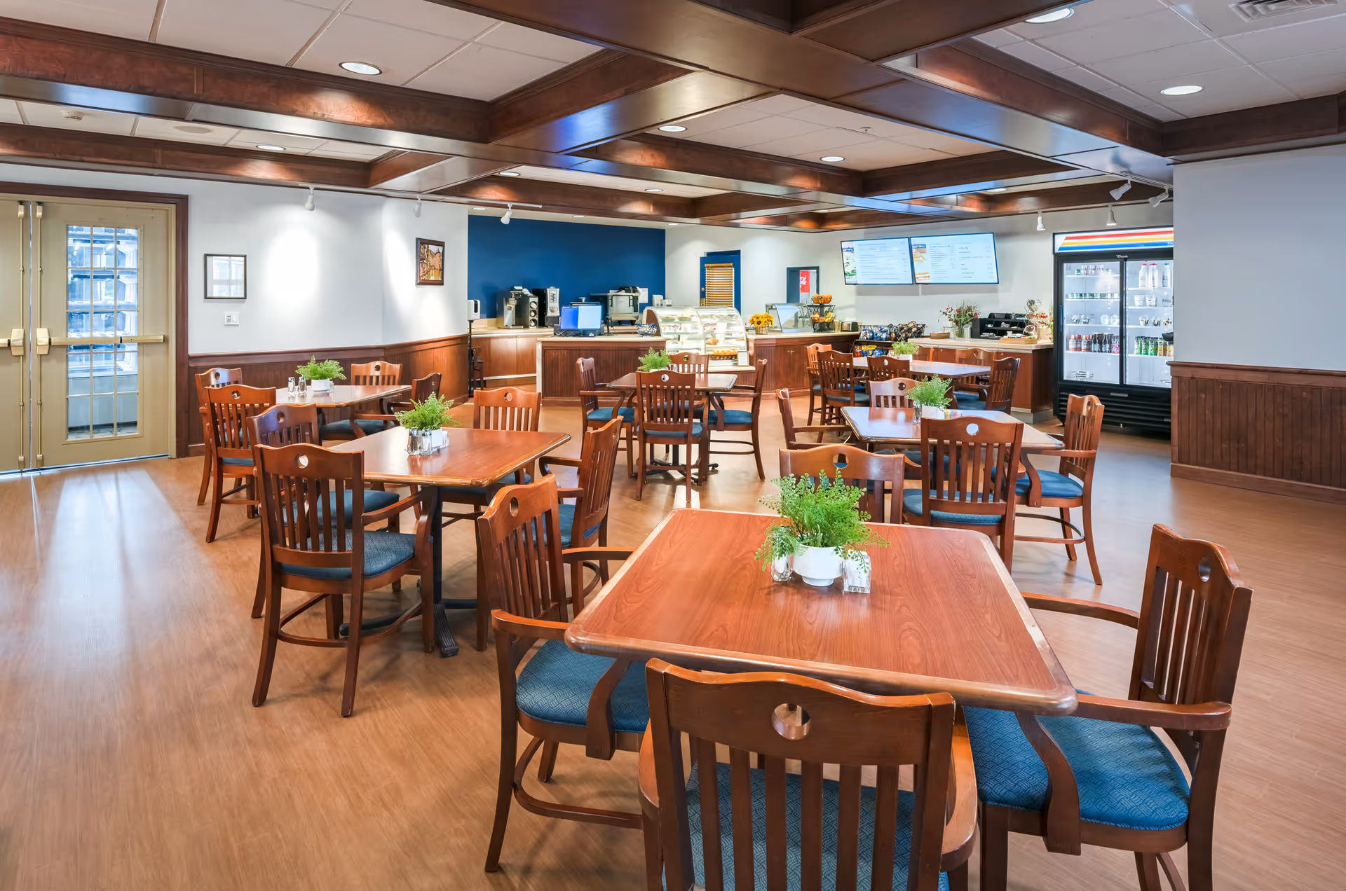 Bright communal dining room with wooden tables and chairs, small potted plants on tables, and a service counter and refrigerated display in the background.