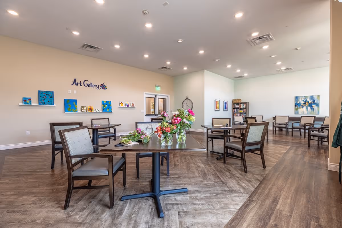 A spacious, well-lit room with wooden flooring and several tables and chairs arranged throughout. The walls feature colorful artwork and a sign that reads 'Art Gallery'. There are vases with fresh flowers on the tables, and a bookshelf with books is visible in the background. The ceiling has multiple recessed lights.