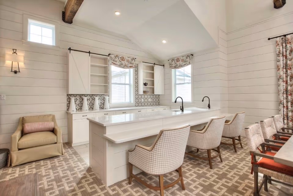 Bright and airy kitchen area with white cabinetry, a large island with two sinks and four cushioned chairs. The walls are white shiplap with patterned tile backsplash and floral curtains on the windows. There is a beige armchair with a pink pillow on the left side and a patterned carpet covering the floor.