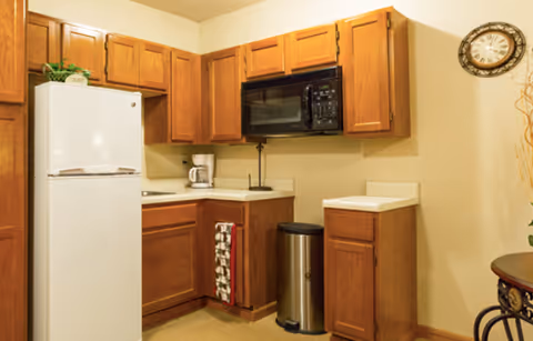 Small kitchenette with wooden cabinets, a white refrigerator, microwave, coffee maker, and a stainless steel trash can.