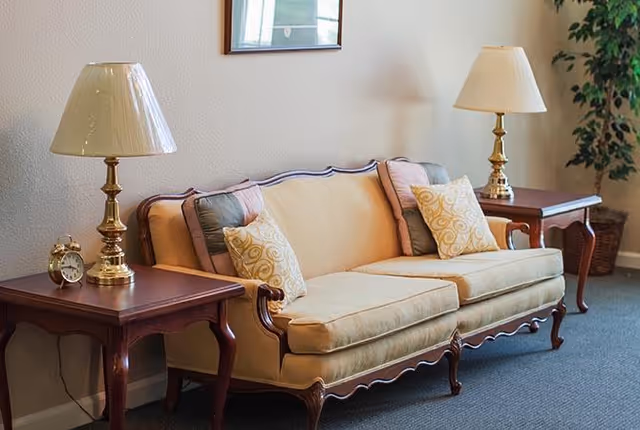 A beige upholstered sofa with wooden frame and decorative pillows placed between two wooden side tables, each with a brass lamp and a small clock on the left table, set against a light-colored wall with a framed picture and a potted plant in the corner.