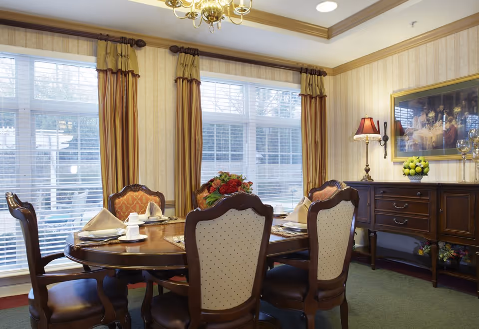 A formal dining room with a round wooden table set for four people with plates, napkins, and cups. The room features large windows with striped curtains, a wooden sideboard with a lamp and a bowl of green apples, and a framed painting on the wall. The walls have striped wallpaper and the floor is carpeted.