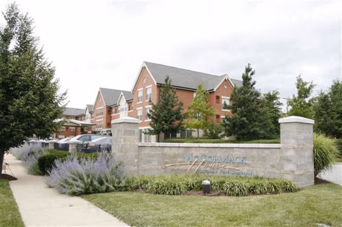 Brick three-story senior living building behind a stone entrance wall and sign, with landscaped shrubs and a sidewalk in front.