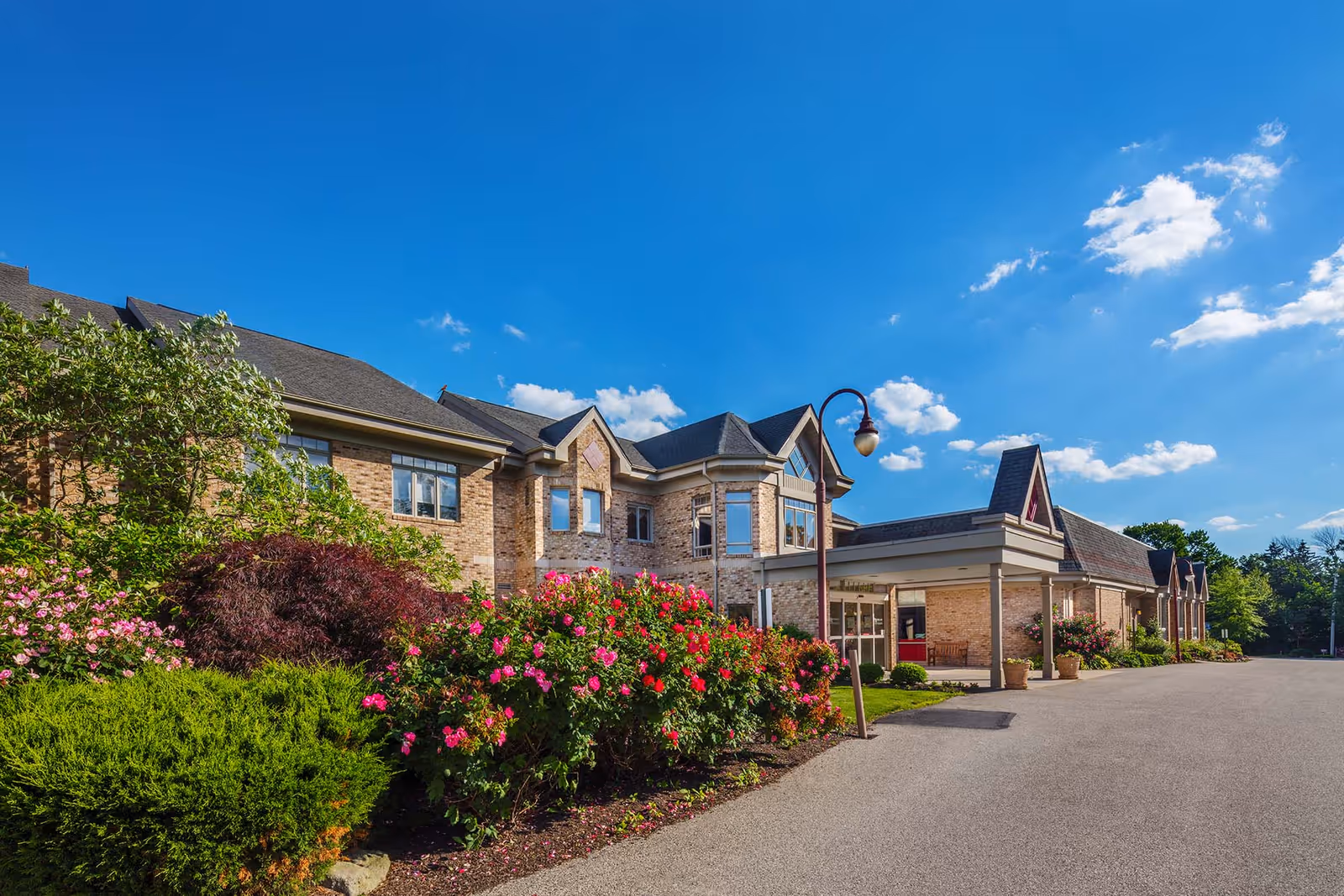 Brick building entrance with colorful flower beds, a lamp post and a paved driveway under a bright blue sky.