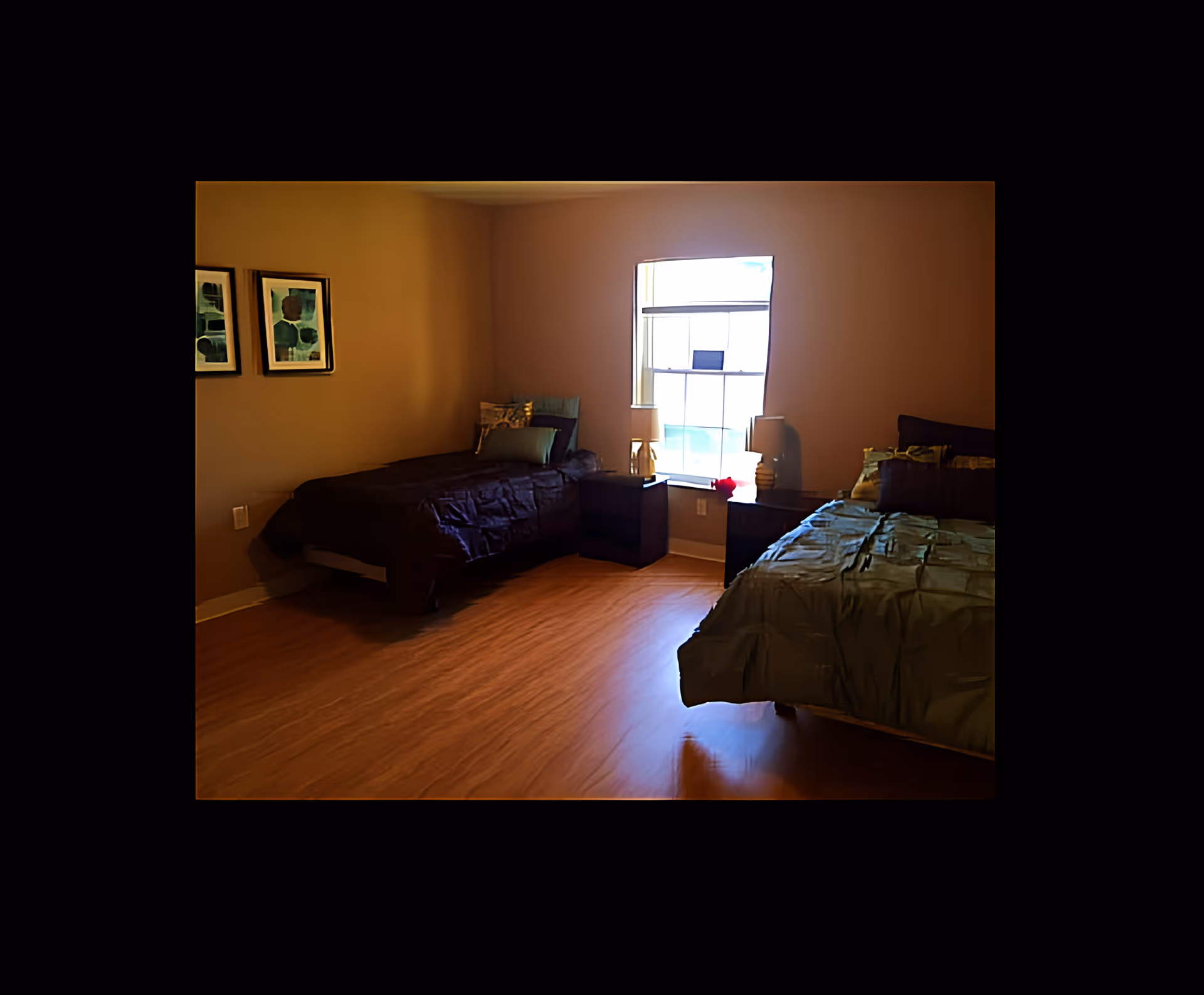 Two-bed bedroom with hardwood floors, a window between nightstands, framed artwork on the wall and warm lighting.