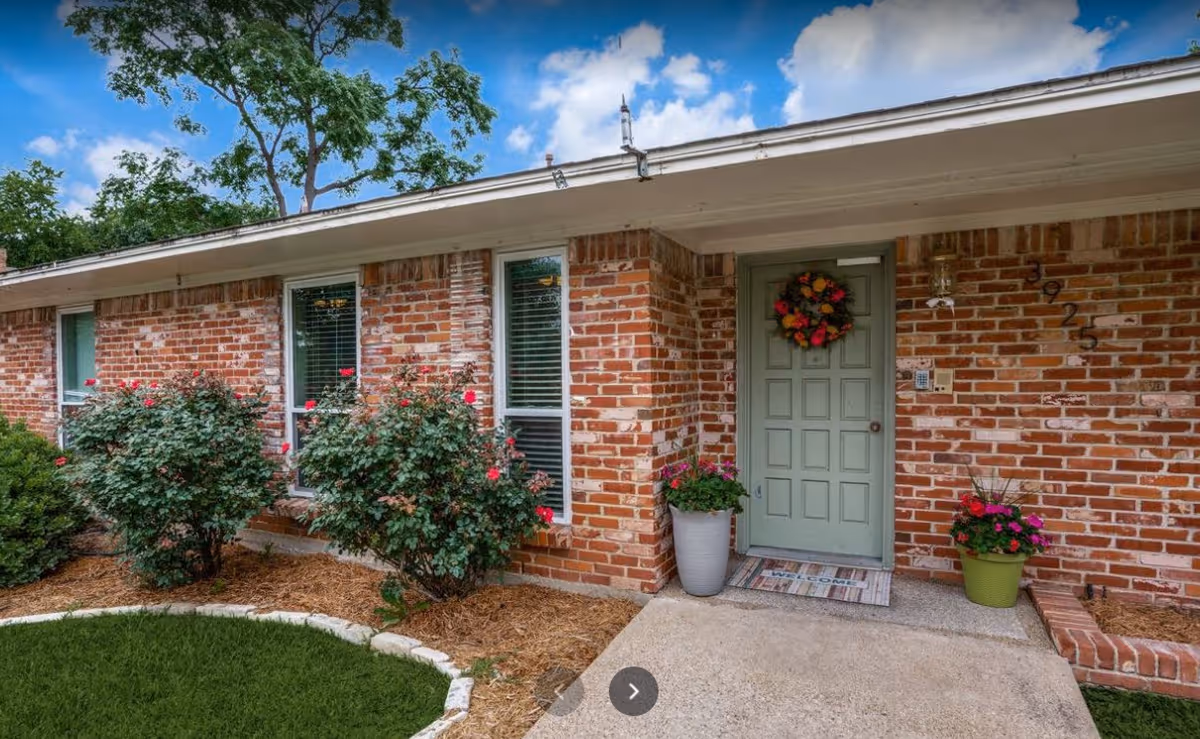Brick single-story home entrance with a pale green front door decorated with a wreath, potted flowers, and rose bushes along the walkway.