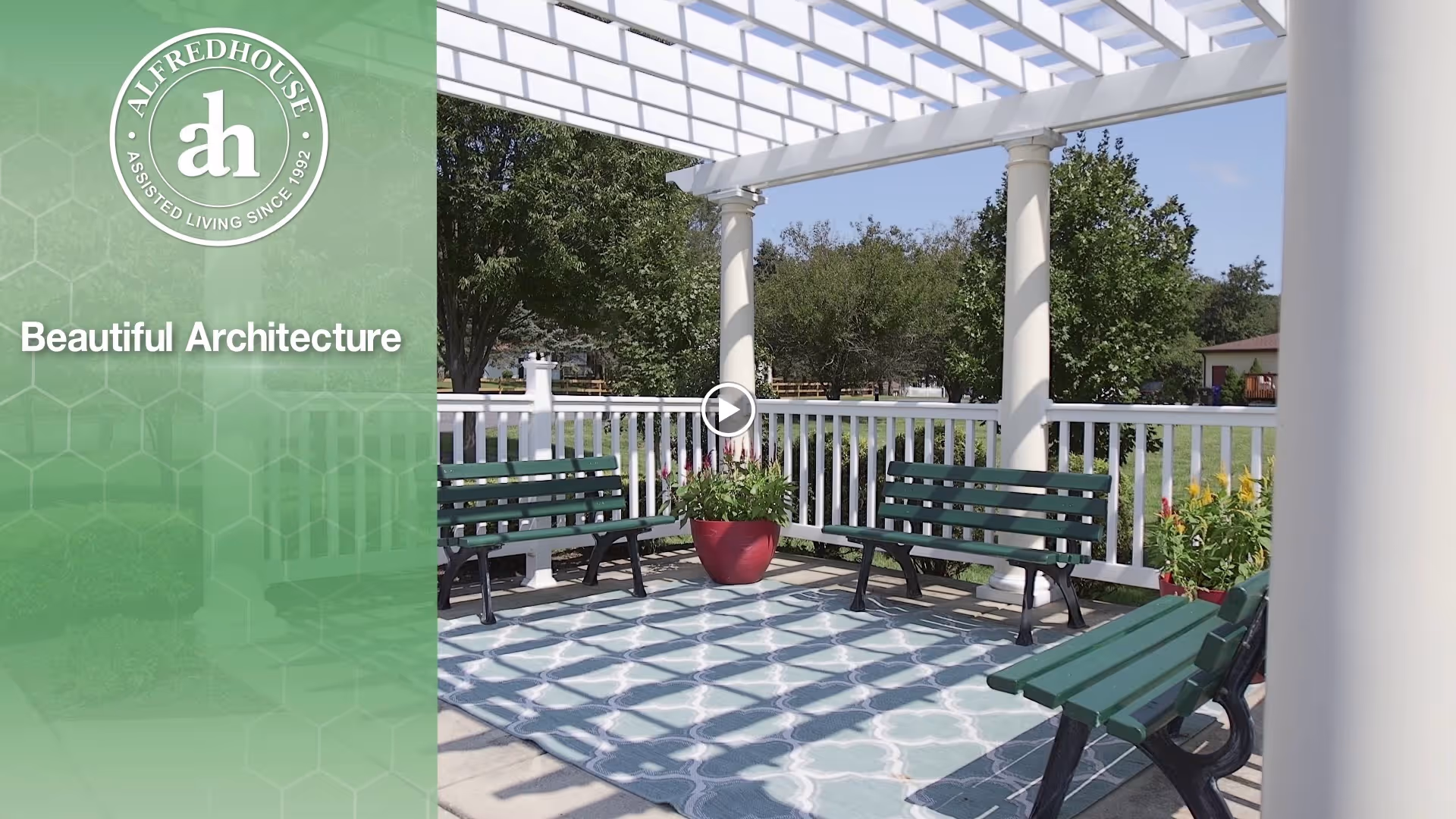 Outdoor seating area with green benches arranged around a patterned outdoor rug under a white pergola. There are potted plants and trees visible in the background, along with a white railing enclosing the space.
