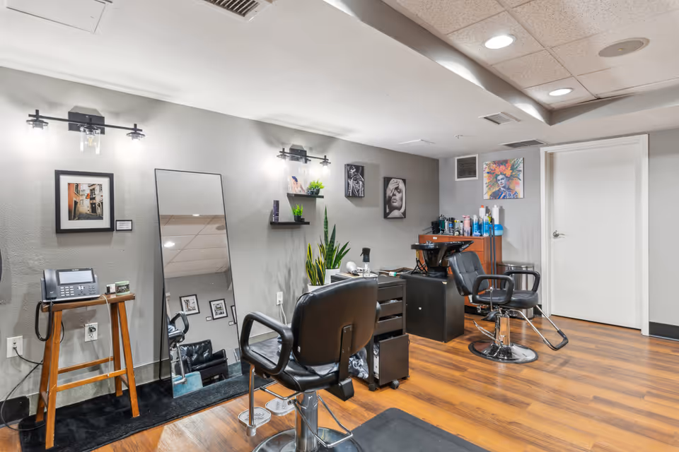 Interior of a hair salon area with two black salon chairs, a large standing mirror, a wooden stool with a telephone on it, shelves with small plants and hair products, and colorful artwork on the walls. The floor is wooden and the ceiling has recessed lighting.