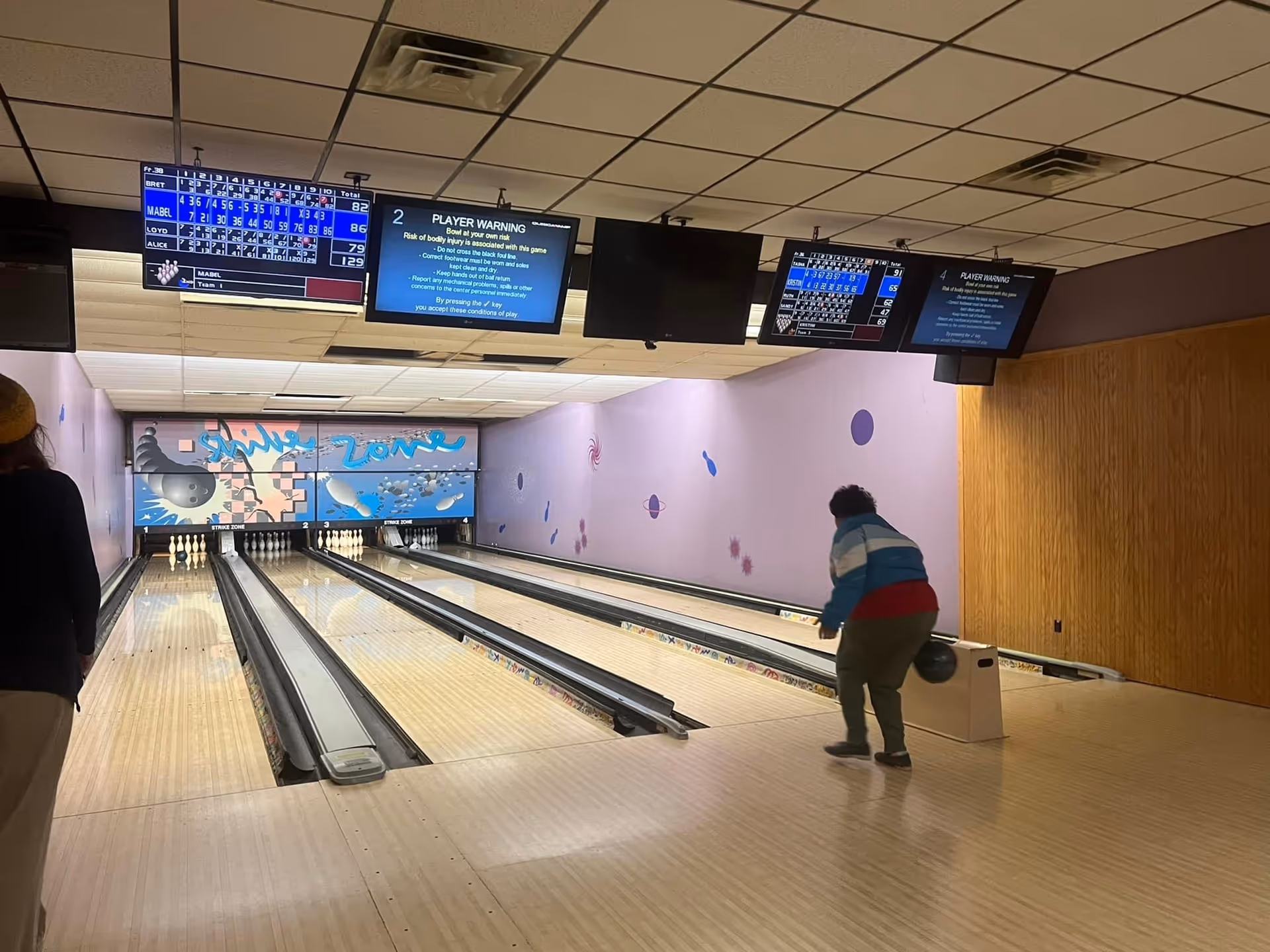 A person rolling a bowling ball down a lane in a small indoor bowling alley with score monitors overhead.
