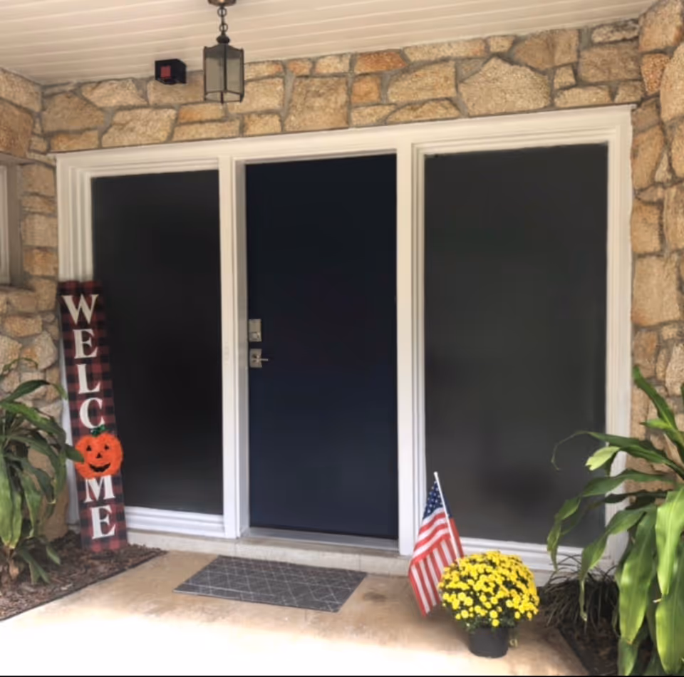 Front entrance of a building with a dark blue door flanked by two large black panels. The surrounding wall is made of stone. There is a hanging lantern light above the door. To the left of the door is a vertical wooden sign with the word 'WELCOME' and a small orange pumpkin decoration. To the right of the door is a pot of yellow flowers with a small American flag placed in it. There are green plants on both sides near the entrance.