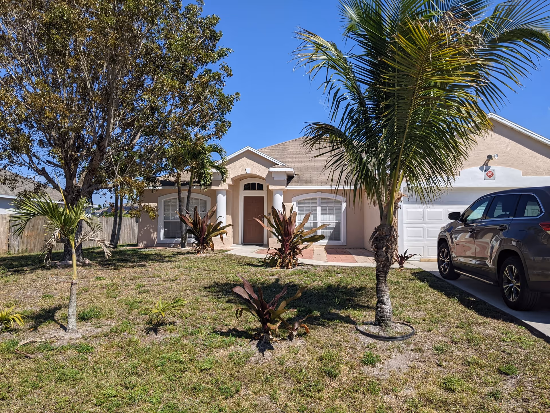 Single-story beige house with an arched front door, garage, front lawn and palm trees under a clear blue sky.