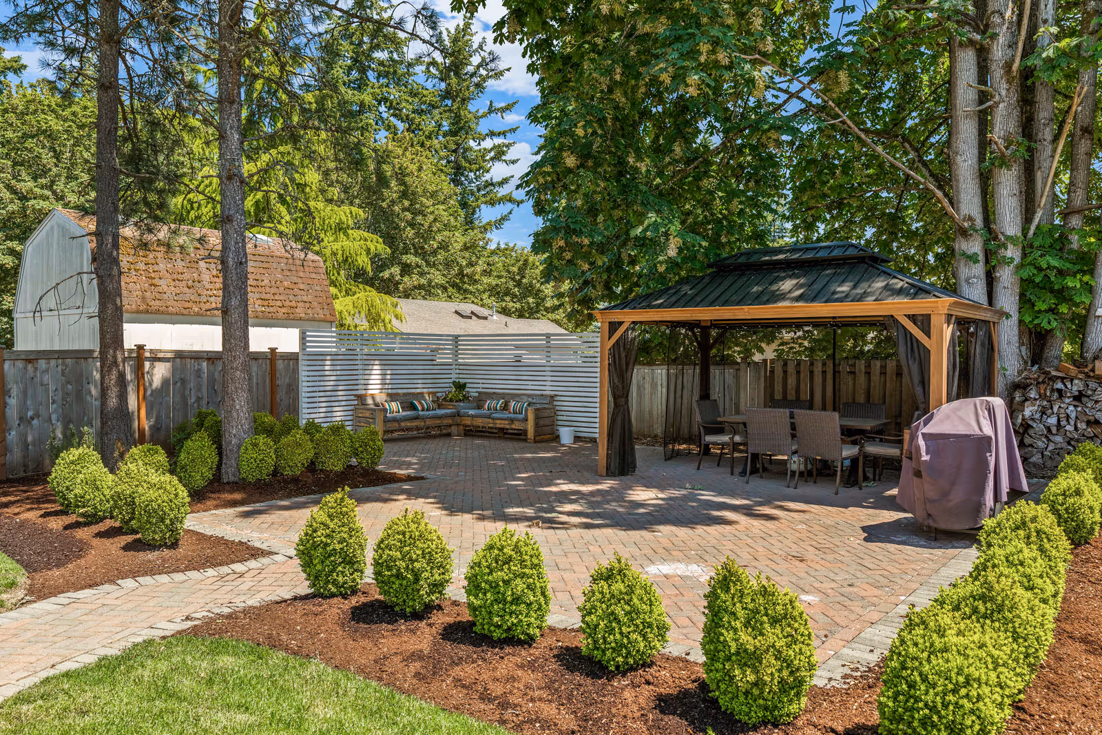 A well-maintained outdoor patio area with a brick-paved ground, a wooden gazebo with a metal roof covering a dining table and chairs, a covered grill, neatly trimmed bushes, tall trees providing shade, and a white wooden fence with a built-in bench adorned with colorful cushions.