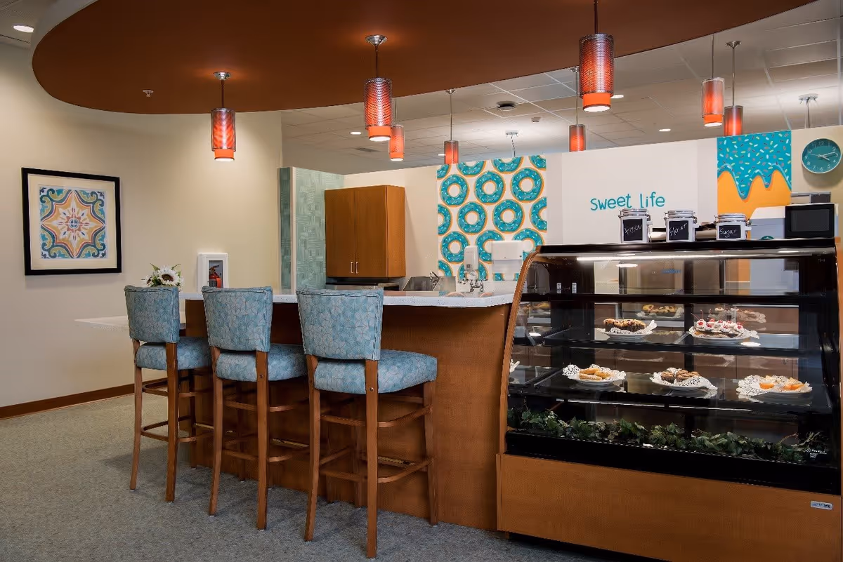 Interior view of a cozy dining area with a counter and four high chairs upholstered in blue fabric. Behind the counter is a display case filled with various pastries and desserts. The wall behind features colorful donut-themed artwork and the phrase 'sweet life'. Several pendant lights hang from the ceiling, illuminating the space warmly.