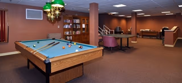 A spacious recreational room with a pool table in the foreground, a bookshelf filled with books, a table tennis table, and several chairs. The room has brown walls and carpeted flooring with ceiling lights providing illumination.