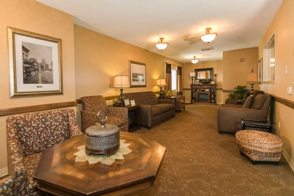 A cozy senior living facility common area with warm beige walls and carpeted floor. The room features multiple seating options including patterned armchairs, a sofa, and a loveseat arranged around wooden side tables with lamps. At the far end, there is a decorative fireplace with a mirror above it and framed pictures on the mantel. The room is softly lit by ceiling lights and table lamps, creating a welcoming atmosphere.
