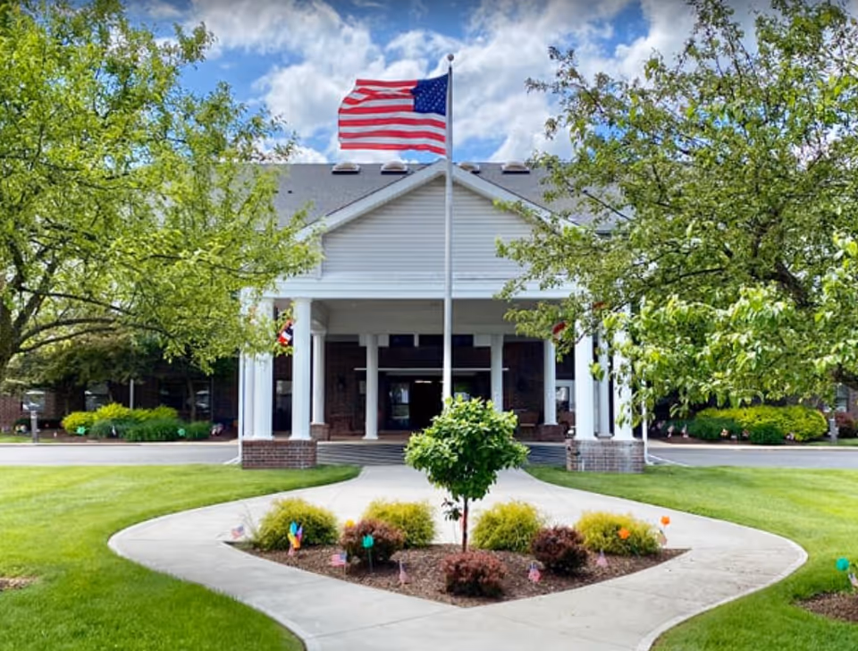 Front exterior view of a building with a covered entrance supported by white columns. A flagpole with an American flag is centered in front of the entrance, surrounded by a landscaped area with small bushes and a tree. Green grass and trees frame the walkway leading to the entrance under a partly cloudy sky.
