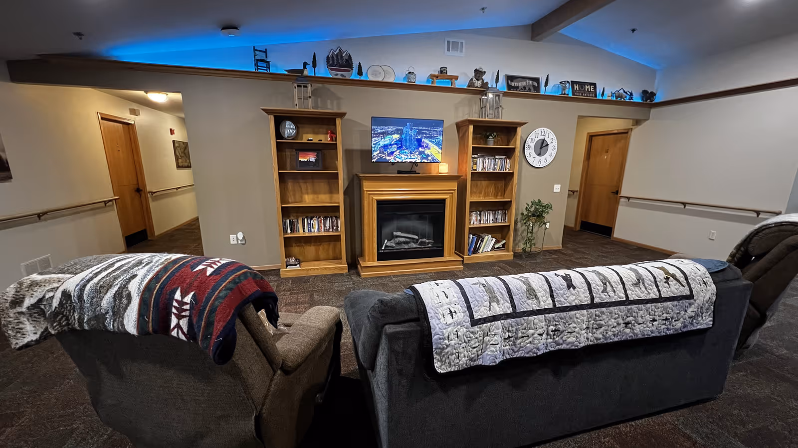 Two sofas with patterned throws face a fireplace and TV set between wooden bookshelves in a common lounge area.