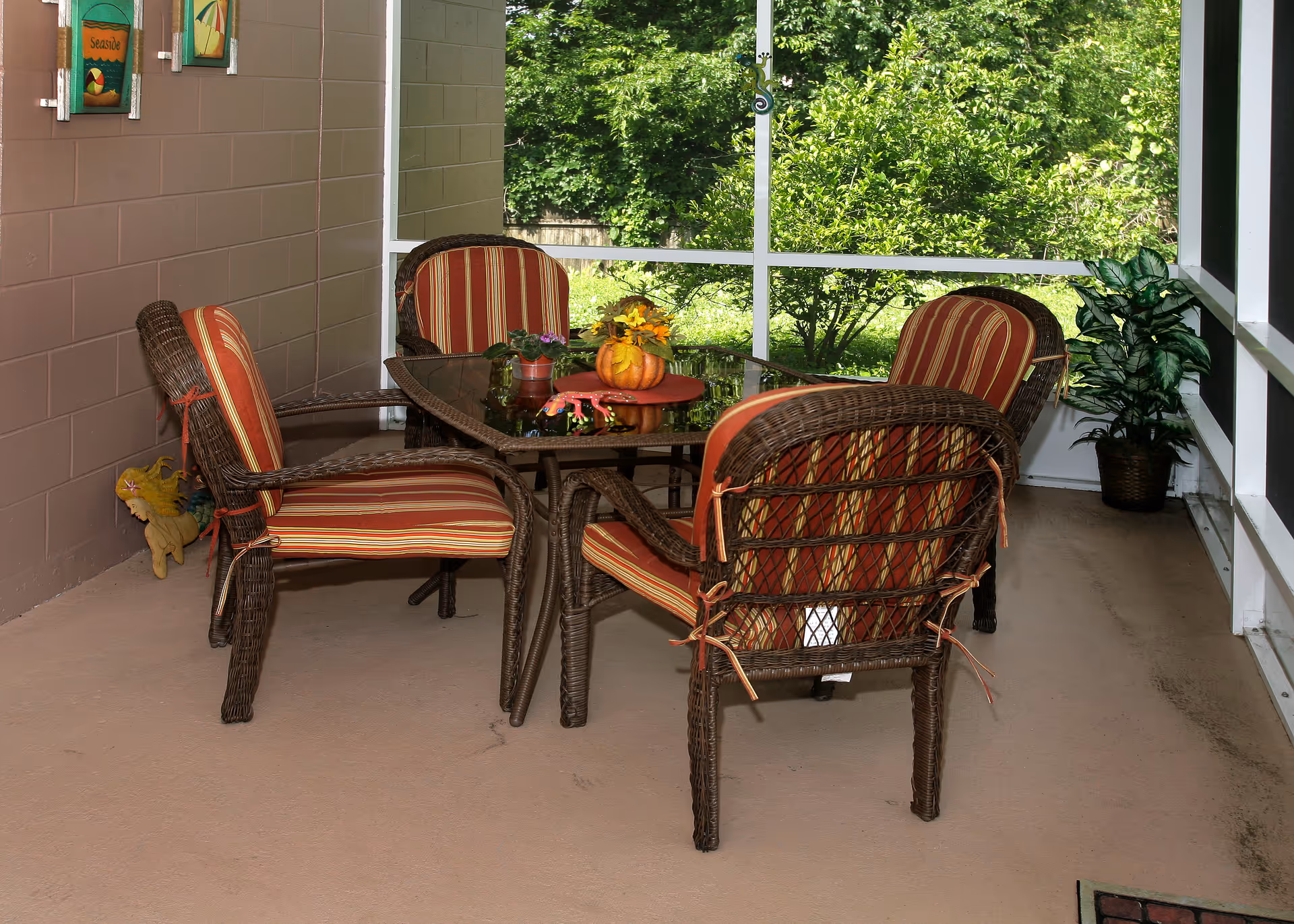 A screened-in porch area with a glass-top table surrounded by four wicker chairs with red and yellow striped cushions. The porch has a view of green trees and bushes outside. There are two small potted plants on the table, one with flowers and one with a small pumpkin decoration. The wall on the left has two framed seaside-themed pictures, and there is a potted plant in the corner on the right side of the porch.
