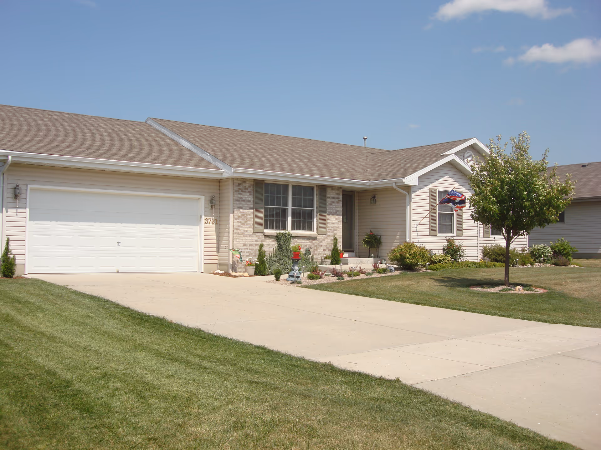 Single-story residential building with a beige exterior, a two-car garage, a small front porch, and a well-maintained lawn with a tree and shrubs under a clear blue sky.