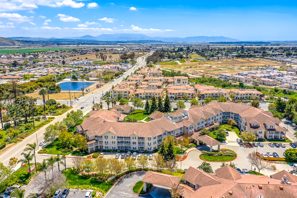 Aerial view of a large assisted living facility with multiple connected buildings featuring red-tiled roofs, surrounded by landscaped greenery, parking lots, and nearby residential neighborhoods under a blue sky with scattered clouds.